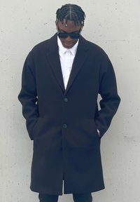 Μαύρο παλτό με δομημένο σχέδιο, που διαθέτει δύο κουμπιά, πλαϊνές τσέπες και λευκό πουκάμισο από κάτω. Λείο υφάσμα.