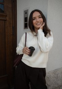 Mladá žena v bílém svetru držící hrnek kávy, usmívající se, stojící proti světlé zdi poblíž dřevěných dveří a interkomu.