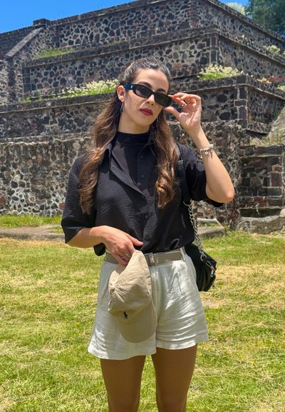 Camisa de botón negra de manga corta, pantalones cortos beige, gorra de béisbol color marrón claro con logo y gafas de sol oscuras, con fondo de una estructura de piedra.