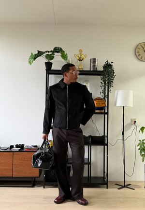 Hombre con chaqueta negra y pantalones marrones sosteniendo una bolsa negra, de pie en interiores cerca de estanterías con plantas, un trofeo y un baloncesto.