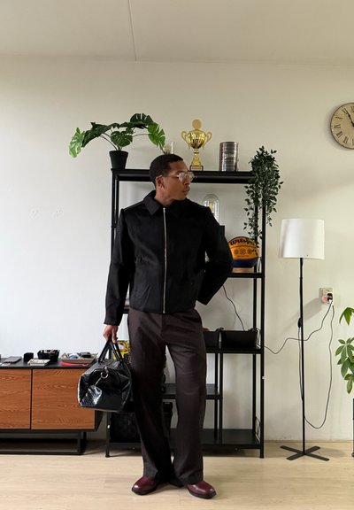 Hombre con chaqueta negra y pantalones marrones sosteniendo una bolsa negra, de pie en interiores cerca de estanterías con plantas, un trofeo y un baloncesto.