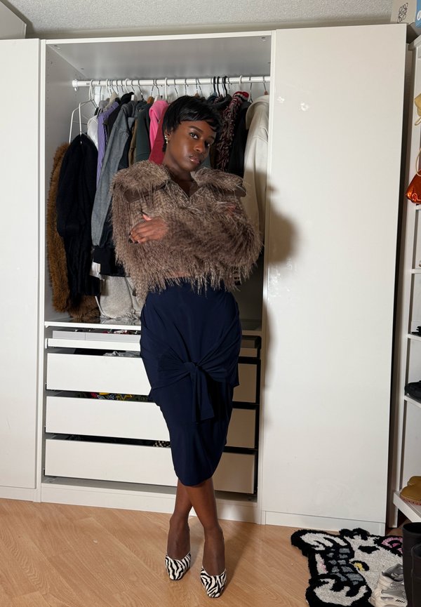 Personne portant une veste en fausse fourrure marron, une jupe bleu marine et des talons à motif zèbre, debout les bras croisés devant une armoire ouverte avec des vêtements.