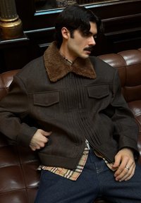 Veste marron à fermeture éclair avec un col en fausse fourrure, présentant des plis horizontaux et deux poches avant. Portée sur une chemise à carreaux et un jean en denim.