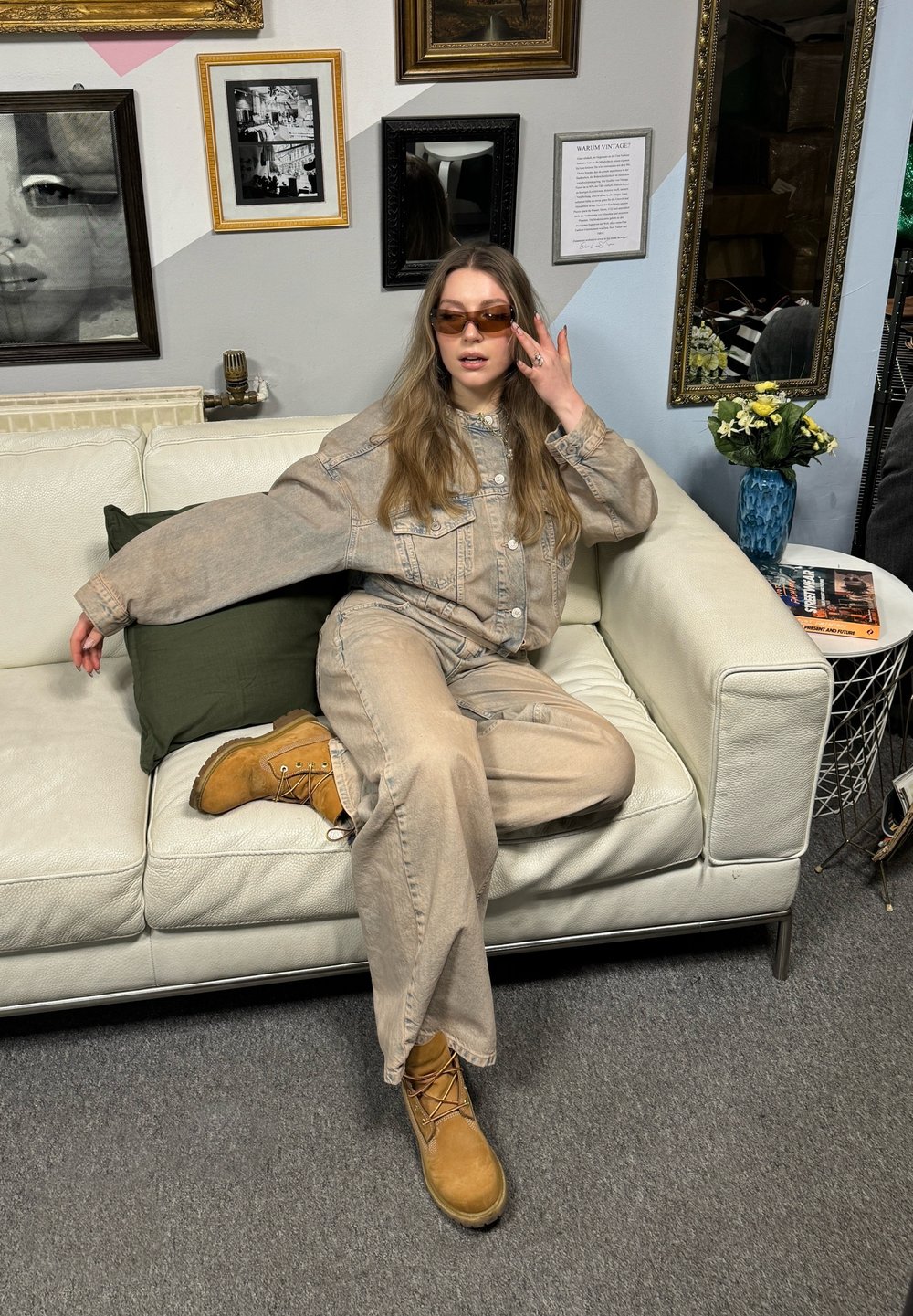Beige denimoutfit med oversized jacka och lösa byxor, tillsammans med bruna arbetsstövlar. Modellen bär bruna solglasögon och sitter på en vit soffa.
