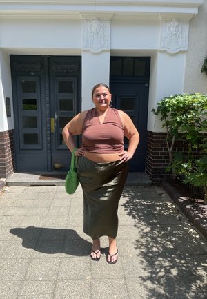 Top sin mangas marrón, falda de satén verde oliva, bolsa de punto verde y chanclas negras, frente a la entrada de un edificio con elementos decorativos.