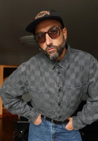 Mann med grå skjegg som bruker solbriller, svart caps, rutete grå skjorte og blå jeans står innendørs med hendene på hoftene.
