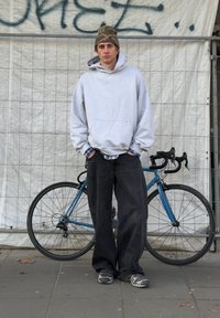 Sweat à capuche gris avec poche avant, chemise à carreaux bleue en dessous, jean noir à jambes larges et baskets grises. Vélo bleu en arrière-plan.