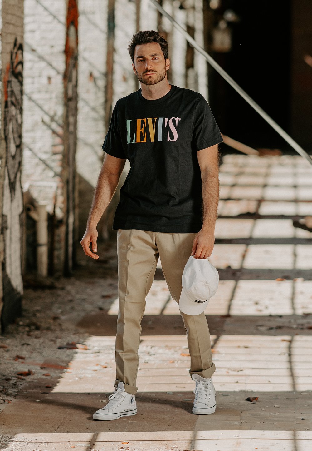Fekete póló színes "LEVI'S" logóval, bézs nadrág, fehér sapka és fehér sportcipő. Átlagos, modern öltözet városi környezetben.