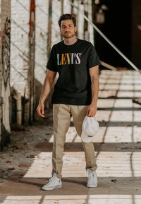 Fekete póló színes "LEVI'S" logóval, bézs nadrág, fehér sapka és fehér sportcipő. Átlagos, modern öltözet városi környezetben.