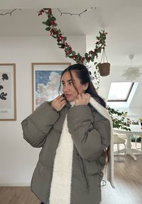 Doudoune vert olive avec un col en fausse fourrure blanche et une fermeture éclair. Silhouette ample avec des détails cousus et une texture douce.