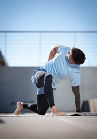 Atletyczny mężczyzna wykonujący rozciąganie w jasno niebieskiej koszulce z krótkim rękawem i czarnych legginsach, boso na macie, na tle czystego niebieskiego nieba.