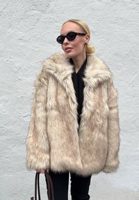 Valepuuskar, hele beeži värvi, paksu tekstuuriga, üleõlakujuline ja laia kraega. Kaasas mustad päikeseprillid ja pruun käekott.