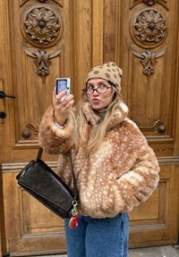 Veste en fausse fourrure marron tachetée ; pull beige ; bonnet marron à motifs ; sac bandoulière sombre avec l'inscription « EXPATRIE » ; porte en bois en arrière-plan.