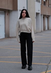 Maglione a trecce color crema con un design cropped, abbinato a pantaloni neri a vita alta. Scarpe nere chunky con plateau completano l'outfit.