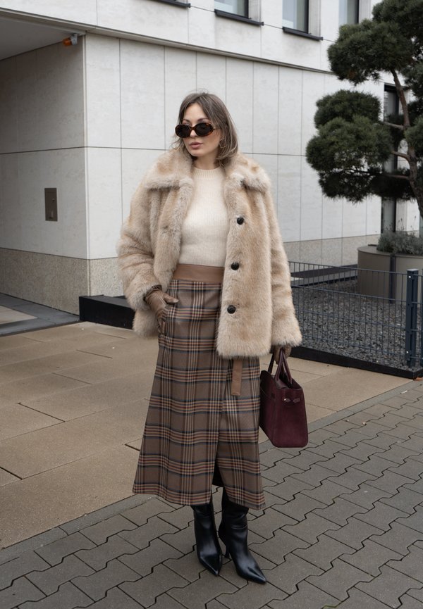 Fuskepels beige frakke, cremefarvet sweater og brun ternet midi nederdel. Sorte spidse støvler og bordeaux håndtaske fuldender looket. Urban omgivelser.