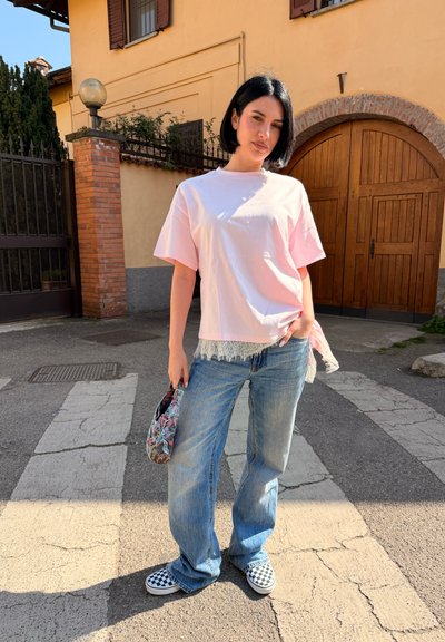 Mujer con pelo corto negro estilo bob, lleva una camiseta rosa claro de gran tamaño, vaqueros azules holgados, zapatos slip-on a cuadros y sostiene un bolso con estampado.