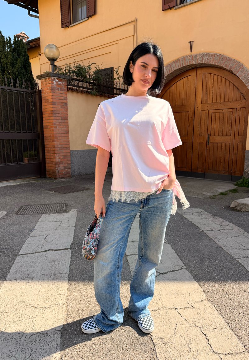 Woman with black bob hair wearing a light pink oversized T-shirt, loose blue jeans, checkered slip-on shoes, and holding a patterned handbag.