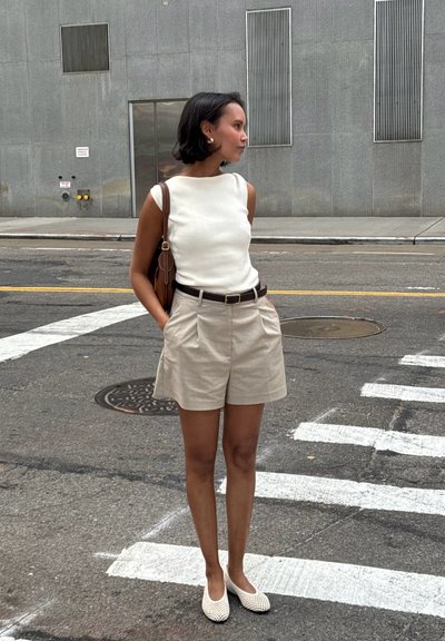 Top blanco sin mangas, pantalones cortos plisados beige, cinturón marrón y mocasines blancos. Bolso marrón colgado del hombro. Fondo urbano.
