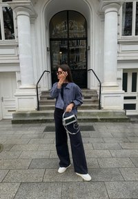 Prúžkovaná modro-biela skrátená košeľa na gombíky, čierne široké nohavice, biele tenisovky, drží priesvitnú kabelku, stojí na kamenných schodoch.