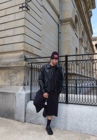 Hombre con chaqueta de cuero negra, falda larga negra y gorro se apoya contra un saliente de piedra con una valla de metal y la fachada de un edificio histórico detrás.