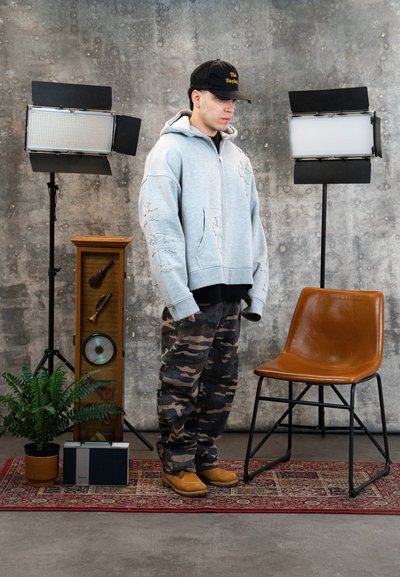 Joven con pantalones de camuflaje, sudadera con capucha gris y gorra negra de pie junto a una silla marrón, un altavoz vintage y dos luces de estudio sobre una alfombra con estampado.