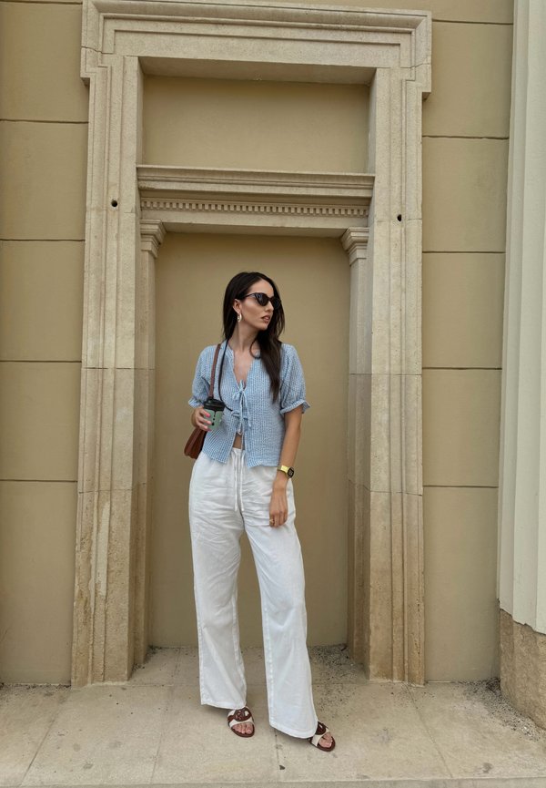 Camicia a maniche corte a scacchi blu, pantaloni bianchi a gamba larga, sandali marroni e occhiali da sole. Tiene una tazza di caffè. Si trova in un'arcata di pietra.