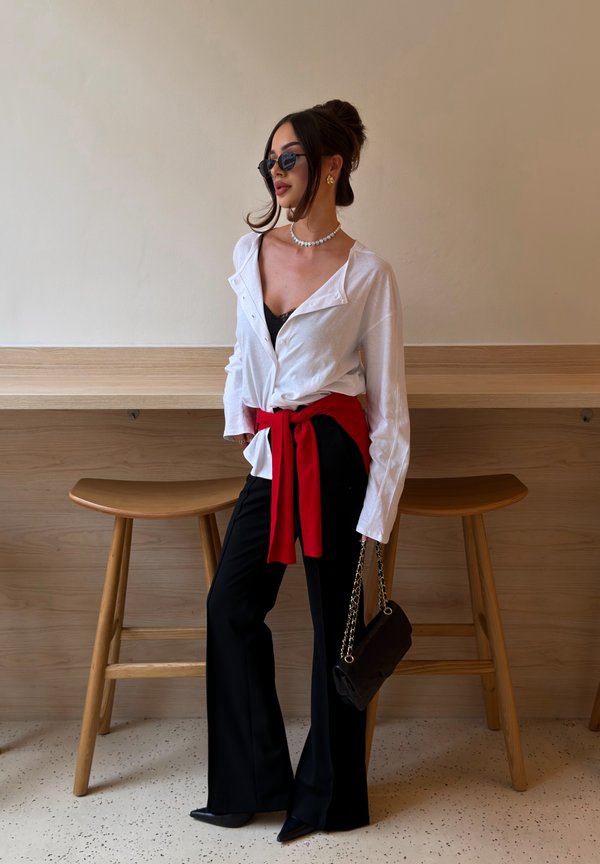 Mujer con camisa blanca sobre top negro, pantalones negros, suéter rojo atado a la cintura, gafas de sol, collar de perlas, sosteniendo bolso negro, de pie junto a taburetes de madera.