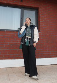 Pantalon noir à jambes larges, chemise à carreaux verts nouée à la taille, sweat à capuche rayé, veste noire et blanche surdimensionnée, baskets grises, cadre extérieur.