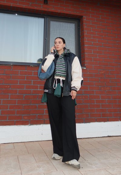 Pantalones anchos negros, camisa de cuadros verde atada a la cintura, sudadera con capucha a rayas, chaqueta grande en blanco y negro, zapatillas grises, ambiente al aire libre.