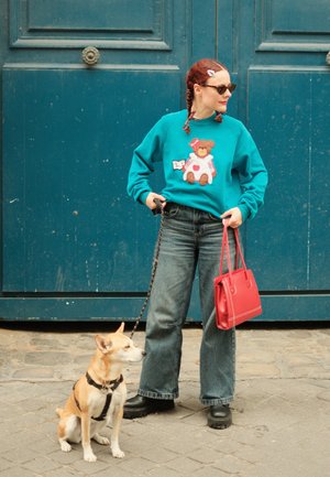 Türkiser Pullover mit Teddybaudesign, hochtaillierte Jeans, schwarze Plateau-Boots und eine rote Tragetasche; ein Hund an der Leine sitzt daneben.