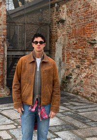 Jeune homme portant une veste en cuir marron, des lunettes de soleil et un jean, debout sur un pavé en pierre devant un mur de briques rustique avec une porte métallique.