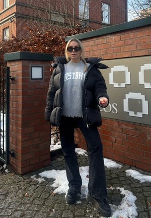 Mujer con gafas de sol, chaqueta acolchada negra, suéter gris y pantalones negros, de pie sobre un empedrado con polvo de nieve cerca de una pared de ladrillo y una puerta.