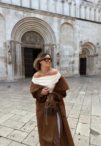 Płaszcz przypominający brązowe futro z szerokimi rękawami, nałożony na jasnokremowy top z odkrytymi ramionami. Mała brązowa torebka jest trzymana na tle kamiennej ściany.