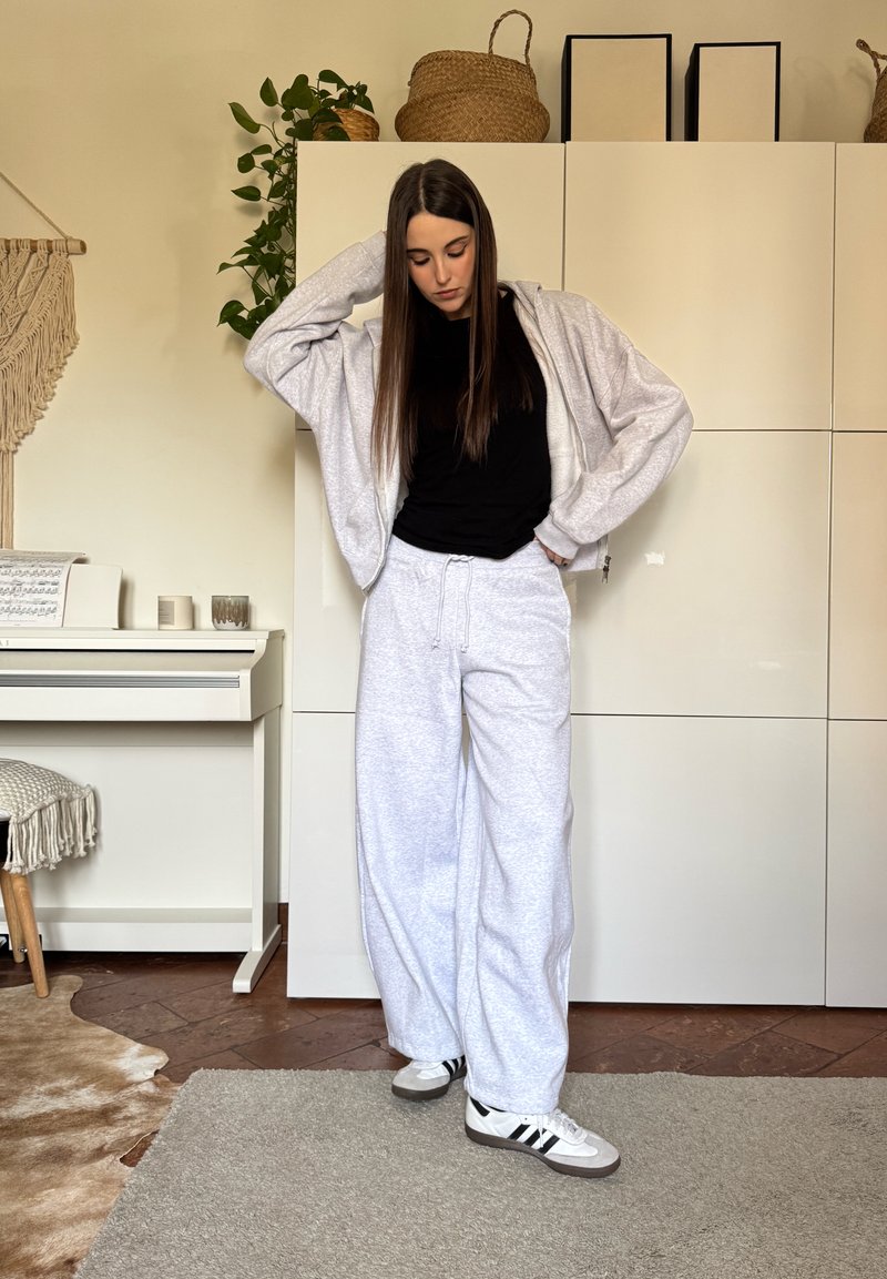 Jeune femme aux longs cheveux, portant un pantalon de survêtement gris, un sweat à capuche gris, un t-shirt noir et des baskets blanches, debout dans une pièce confortable avec des plantes et un piano.