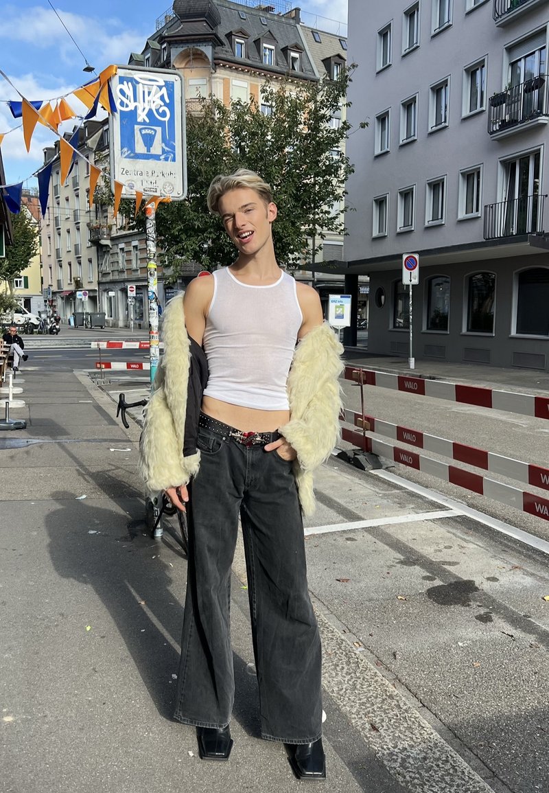 Jeune personne posant avec confiance sur un trottoir en ville, portant un débardeur blanc, un jean large noir, des bottes noires et une veste en fourrure crème.