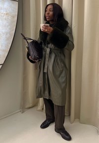 Olivgrüner Leder-Trenchcoat mit Kunstfellkragen, tailliert, kombiniert mit braunen Hosen und schwarzen Slippern. Hält eine Tasse und eine Tasche.