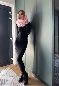 Crna haljina dugih rukava, prilagođenog kroja, uparena s svijetloružičastim šalom. Model stoji uz zelenu kuhinjsku jedinicu, noseći robusne crne čizme.