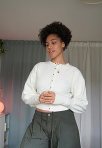 Weißer Strickcardigan mit rundem Ausschnitt und Knopfleiste, kombiniert mit locker sitzenden olivgrünen Hosen. Weiche Textur, lange Ärmel und entspannte Passform.