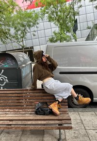 Jeune femme en sweat à capuche marron et jupe grise assise sur un banc en bois à côté d'une camionnette blanche, avec un sac noir à côté d'elle, fond urbain avec graffiti.