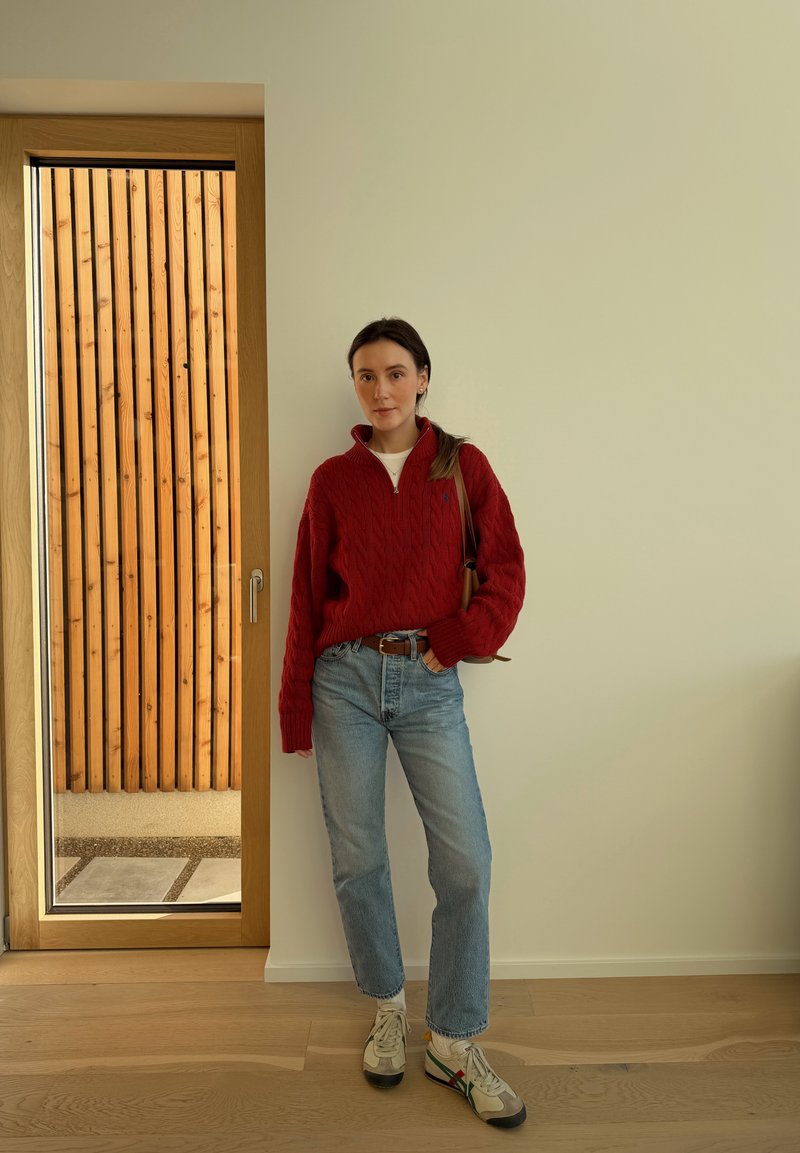 Femme portant un pull rouge, un jean bleu clair et des baskets blanches, debout à l'intérieur contre un mur blanc près d'une porte en verre encadrée de bois.