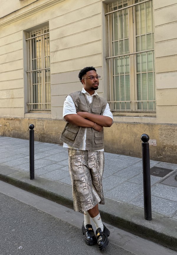 Homme avec des lunettes, en gilet sans manches et pantalon large à motifs, debout sur le trottoir devant un bâtiment beige avec des fenêtres grillagées.