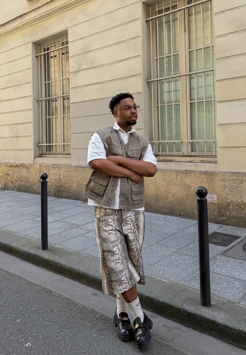 Homme portant des lunettes, un gilet sans manches et un pantalon large à motifs, debout sur le trottoir devant un bâtiment beige avec des fenêtres grillagées.