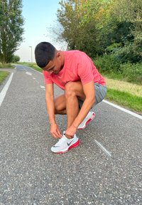 Fehér sportcipők piros részletekkel és fekete swoosh-sal, egy rózsaszín pólóval és szürke rövidnadrággal párosítva, amelyet egy aszfaltúton kötnek meg.
