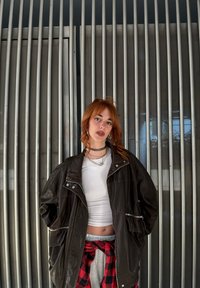 Jachetă oversized din piele maro închis, purtată peste un top alb, cu pantaloni de trening gri și o flanelă în carouri roșii și negre legată în talie.