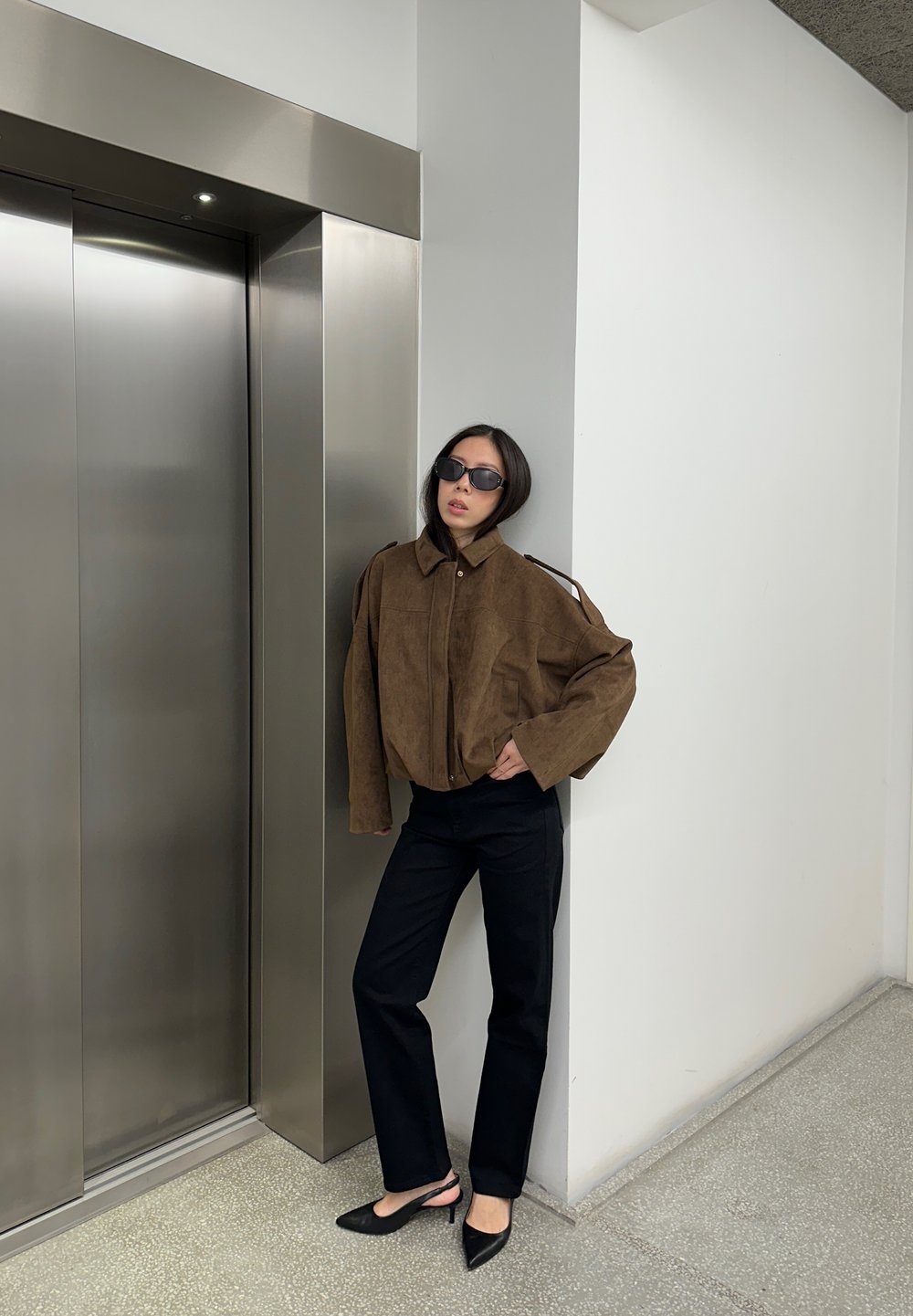 Veste en daim marron avec un col haut, pantalons noirs, et talons noirs pointus ; debout près d'un ascenseur métallique, fond minimaliste.