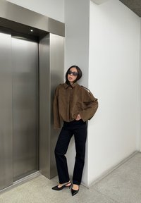 Veste en daim marron avec un col haut, pantalons noirs, et talons noirs pointus ; debout près d'un ascenseur métallique, fond minimaliste.