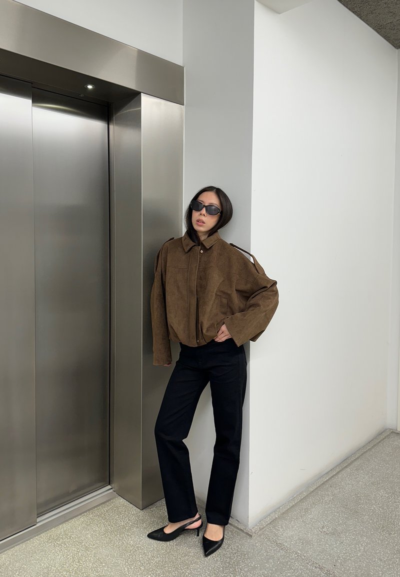 Smeđa jakna od antilopa s visokim ovratnikom, crne hlače i crne šiljaste štikle; stoji pored metalnog lifta, minimalistička pozadina.