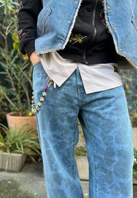 Colete de ganga em azul claro com bordas desgastadas, sobre uma camisola preta e uma camisa cinza, complementado por jeans azuis estampados e um porta-chaves decorativo com contas.