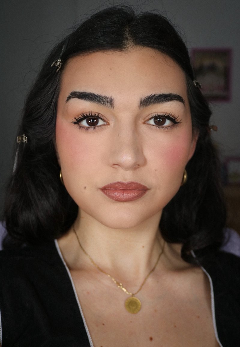 Jeune femme aux cheveux ondulés foncés repliés derrière les oreilles, portant des boucles d’oreilles créoles en or, un collier en or avec un pendentif rond et un maquillage subtil.