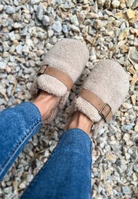 Beiga, mjuka tofflor i fuzzy-typ med en mjuk shearling-textur och ett brunt mockaband med en silverfärgad spänne, bärs tillsammans med blå jeans.
